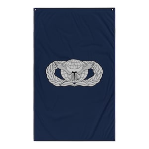 Könnte beinhalten: Eine marineblaue Flagge mit einem silbernen Emblem. Das Emblem zeigt einen Globus mit einem ausgebreiteten Adler, flankiert von Lorbeerkränzen. Die Flagge ist rechteckig und hat oben eine Öse zum Aufhängen.