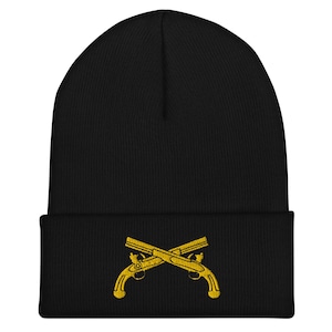 May include: Black beanie with a gold embroidered design of two crossed pistols. The beanie is made of a soft, knitted material and has a folded cuff. The gold embroidery provides a striking contrast against the dark background.