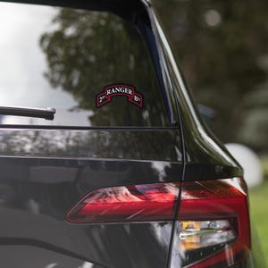 Puede incluir: Una pegatina negra con el texto "2D RANGER BN" en letras rojas y blancas. La pegatina tiene forma de pancarta y está adherida a la ventana trasera de un coche negro.