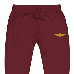 May include: Burgundy sweatpants with a drawstring waist and side pockets. A gold-colored embroidered emblem of wings and a circle is on the left leg. The pants are made of a soft, comfortable material.