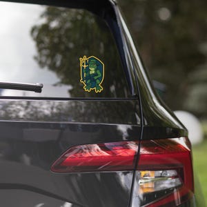 May include: A sticker featuring a green frog character holding a trident and wearing a hat, framed by a yellow and blue shield. The sticker is on the rear window of a dark car.