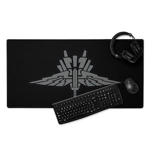 Pode incluir: Tapete de mesa retangular preto com um emblema cinza, um teclado, um rato e auscultadores pretos. O emblema é um design estilizado com asas e um elemento central em forma de espada. O teclado é preto com letras brancas.