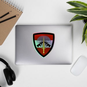 May include: A red, white, and blue shield-shaped sticker with a parachute, swords, and guns. The sticker has a holographic effect.