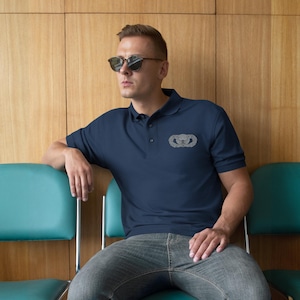 Könnte beinhalten: Marineblaues Poloshirt mit silbernem Emblem auf der linken Brust. Das Shirt hat einen klassischen Kragen und eine Knopfleiste. Die Person auf dem Bild trägt eine Sonnenbrille und Jeans.