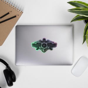 May include: A black and iridescent sticker with a detailed emblem design, including a star, anchor, and crossed weapons. The sticker, with a metallic sheen, is placed on a laptop. The edges of the sticker have a subtle rainbow effect.