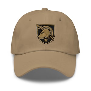 Pode incluir: Boné de beisebol bege com aba curva. O boné apresenta um emblema bordado de um capacete, espada e estrela em dourado e preto. O boné é feito de tecido macio e tem uma alça ajustável.