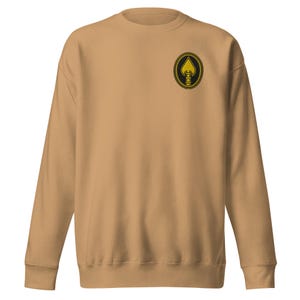 Op de afbeelding: Een bruin sweatshirt met een ronde hals en lange mouwen. Een cirkelvormige patch op de linkerbovenborst heeft een gouden schoppenontwerp binnen een zwart-gouden touwrand. De sweatshirt is gemaakt van zacht materiaal.