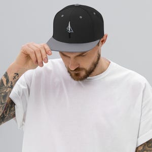 May include: Black and gray baseball cap with a flat brim. The cap features a white and gray arrow design on the front. The cap is being held by a person.