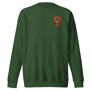 May include: A dark green sweatshirt with a small embroidered patch on the upper left chest. The patch features a skull, wings, and other military-style insignia in red, yellow, and black.