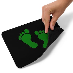 Può includere: Tappetino rettangolare nero con due impronte verdi. Una mano solleva il tappetino, rivelando un lato inferiore strutturato. Le impronte sono di un verde brillante e uniforme.