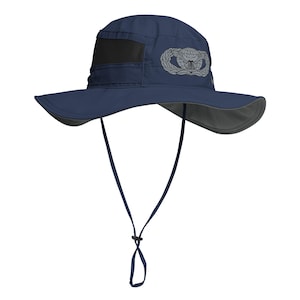 May include: Navy blue sun hat with a wide brim and a black mesh panel. Features a silver emblem on the side and a chin strap. The hat is designed for sun protection and outdoor activities.
