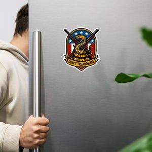 May include: A colorful sticker featuring a coiled rattlesnake, crossed rifles, and the text "DON'T TREAD ON ME." The design incorporates red, white, and blue elements, with a yellow banner. The sticker is affixed to a metallic surface.