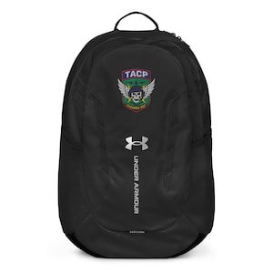 Pode incluir: Mochila preta Under Armour com um logotipo TACP com uma caveira, asas e o texto "CLEARED HOT". A mochila tem uma alça superior, um bolso lateral de malha e o logotipo Under Armour em branco. Projetada para transportar itens.