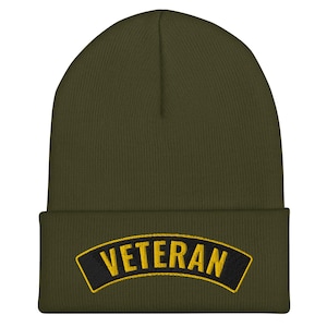 May include: An olive green beanie hat with a black and gold embroidered "VETERAN" patch. The hat is made of a soft, knitted material. A classic style, suitable for cold weather. The hat is a dark green colour.