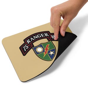 Könnte beinhalten: Beigefarbenes Mauspad mit dem Emblem des 75. Ranger Regiments. Das Emblem zeigt ein Banner mit der Aufschrift "75 RANGER", ein Schild mit Sonne, Blitz und Stern. Das Mauspad hat eine schwarze, strukturierte Unterseite.