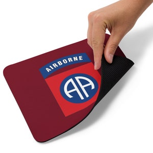 May include: A red mouse pad with a blue and white 101st Airborne Division insignia. The insignia features the letters "AA" inside a circle. The text "AIRBORNE" is above the insignia.