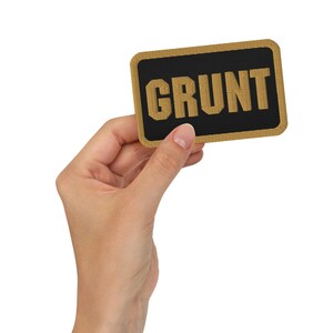 May include: A rectangular patch with the word "GRUNT" embroidered in gold thread on a black background. The patch has a gold border. The patch is held in a person's hand.