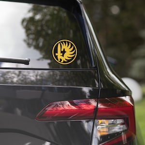 May include: A circular sticker applied to a car window featuring a yellow emblem on a black background. The design depicts a stylized hand holding a vertical sword, surrounded by a curved, wing-like shape, representing the French Foreign Legion.