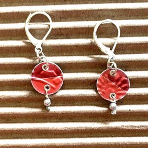 May include: A pair of handmade earrings with red circular pendants and silver lever-back closures. Each earring has a small silver bead hanging from the bottom. The earrings are displayed on a textured brown surface.