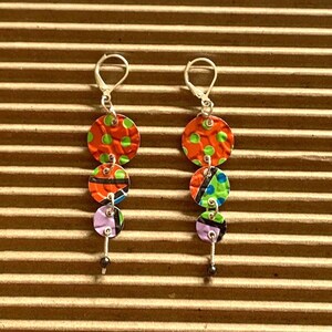 May include: A pair of handmade dangle earrings. Each earring has three colourful, textured circles: orange with green spots, a circle with black, orange and blue designs, and a purple circle. They have silver hooks and a small black bead at the bottom.