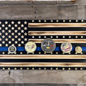 May include: A wooden American flag with a blue stripe, featuring a black background and a field of stars. The flag has a distressed wood finish and is decorated with military challenge coins. The coins are arranged in a row across the blue stripe, with a thin blue line running through the center of the stripe.