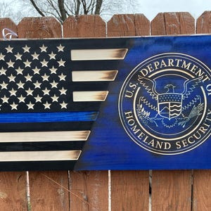 May include: A wooden American flag with a blue line and the U.S. Department of Homeland Security seal. The flag is painted in black, white, and blue.