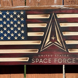 May include: Wooden American flag with the United States Space Force logo. The flag features a blue field with white stars, red and white stripes, and the Space Force emblem. The words "UNITED STATES SPACE FORCE" are below the emblem.