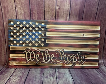 May include: A wooden American flag with a blue and white star field and red and white stripes. The words "We the People" are carved into the wood below the flag.