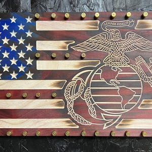 May include: A wooden American flag with the Marine Corps emblem in the center. The flag is decorated with brass bullet casings. The emblem features a globe, an anchor, and an eagle with the words "Semper Fidelis" above it.