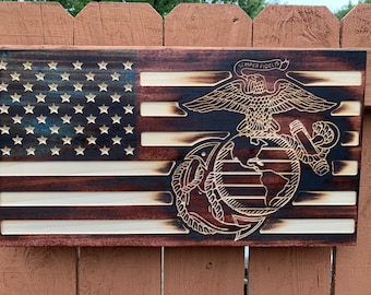 Puede incluir: Bandera americana de madera con el emblema del Cuerpo de Marines. La bandera presenta estrellas y rayas talladas, con el emblema de un águila, un globo terráqueo y un ancla. Las palabras "Semper Fidelis" están sobre el emblema. La madera tiene un tinte oscuro.