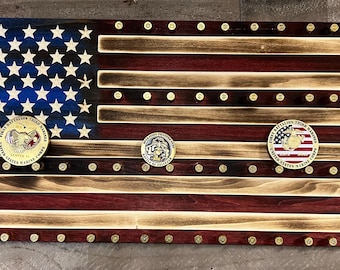 May include: A wooden American flag with a distressed finish, featuring a blue field of stars and red and white stripes. The flag has three gold-colored military challenge coins attached to it. The coins feature the words "First Battalion, Third Marines" and "United States Marine Corps".