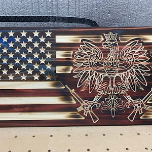 May include: A wooden American flag with a burnt wood finish. The flag is split in half, with the top half showing the blue field of stars and the bottom half showing the red and white stripes. A white eagle with a crown on its head is in the center of the flag, with two rifles crossed behind it.
