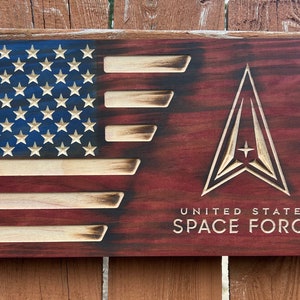 May include: A wooden sign with a distressed, burnt look. The left side features a carved American flag with red, white, and blue colors. The right side features a carved Space Force logo with the text "UNITED STATES SPACE FORCE" below it.