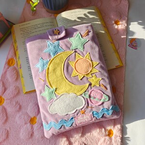 May include: A lavender corduroy book sleeve featuring felt celestial appliques including a large yellow crescent moon, a sun, stars, a cloud, and a planet. The sleeve has a small tab closure with a star button and a blue wavy trim at the bottom.