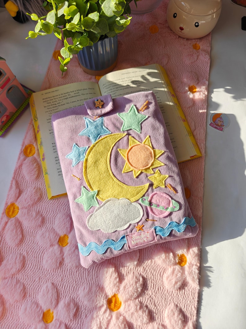 May include: A lavender corduroy book sleeve featuring felt celestial appliques including a large yellow crescent moon, a sun, stars, a cloud, and a planet. The sleeve has a small tab closure with a star button and a blue wavy trim at the bottom.