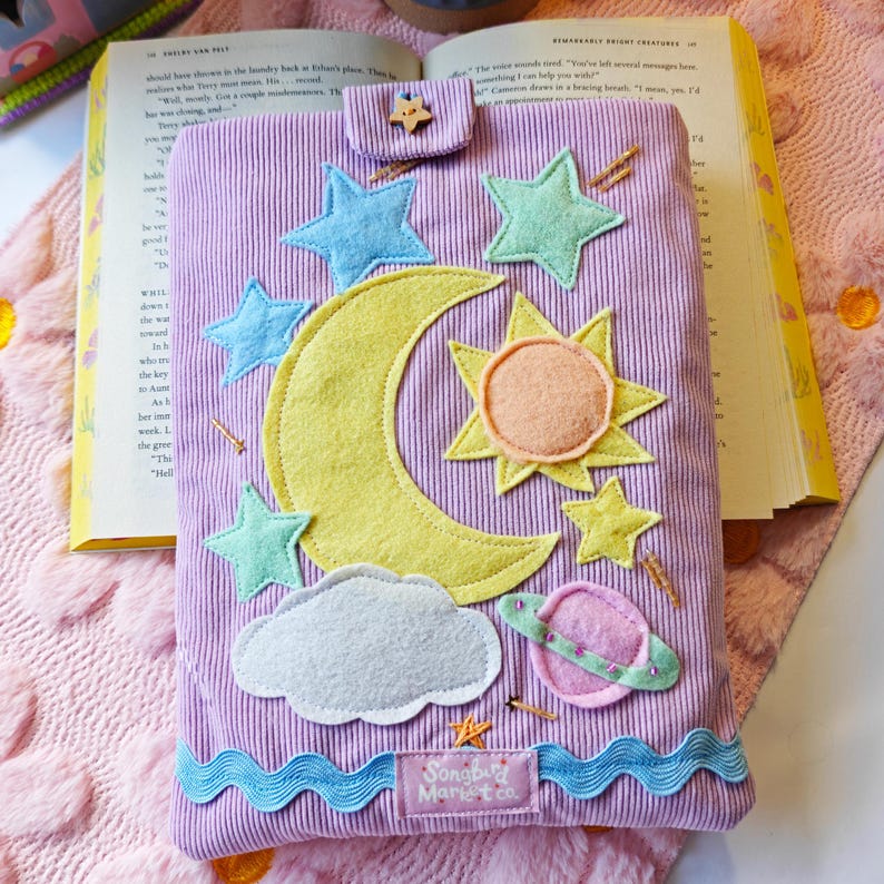 A handmade lavender corduroy book sleeve lies flat on a white surface, resting on a pink floral-patterned rug. The sleeve is decorated with colorful fabric appliqués of a large yellow crescent moon, a yellow and orange sun, a pink ringed planet, and several light blue and green stars. Small, hand-beaded gold details represent shimmering stars scattered across the design, and a wavy blue trim accents the bottom edge. A small wooden star-shaped button secures the top flap.