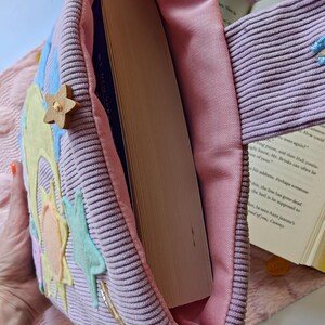 May include: A lavender corduroy tote bag with a pink fabric lining, containing a thick hardcover book. The exterior features felt appliques of a yellow sun, light blue stars, and a small wooden star button attached to the side of the bag.