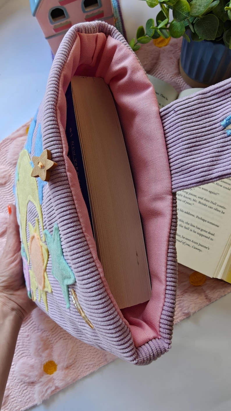 May include: A lavender corduroy tote bag with a pink fabric lining, containing a thick hardcover book. The exterior features felt appliques of a yellow sun, light blue stars, and a small wooden star button attached to the side of the bag.