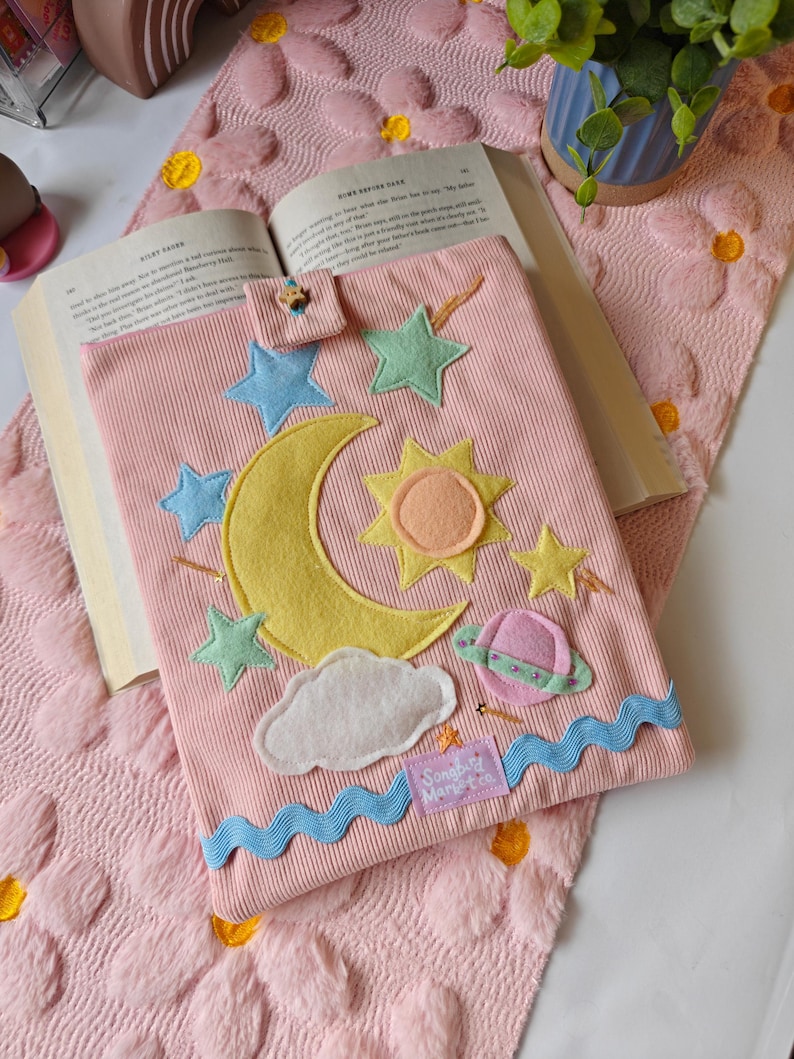 May include: A pink corduroy book sleeve featuring felt celestial appliqu&eacute;s including a yellow crescent moon, a sun, stars, a planet, and a white cloud. The sleeve has a small tab closure with a wooden star button and a blue wavy trim at the bottom.