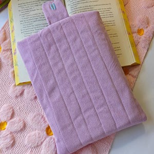 May include: A rectangular lavender corduroy pouch with a vertical ribbed texture and a small matching tab at the top center. The pouch is positioned flat on an open book, which rests on a soft, light pink fabric surface with a raised floral pattern.