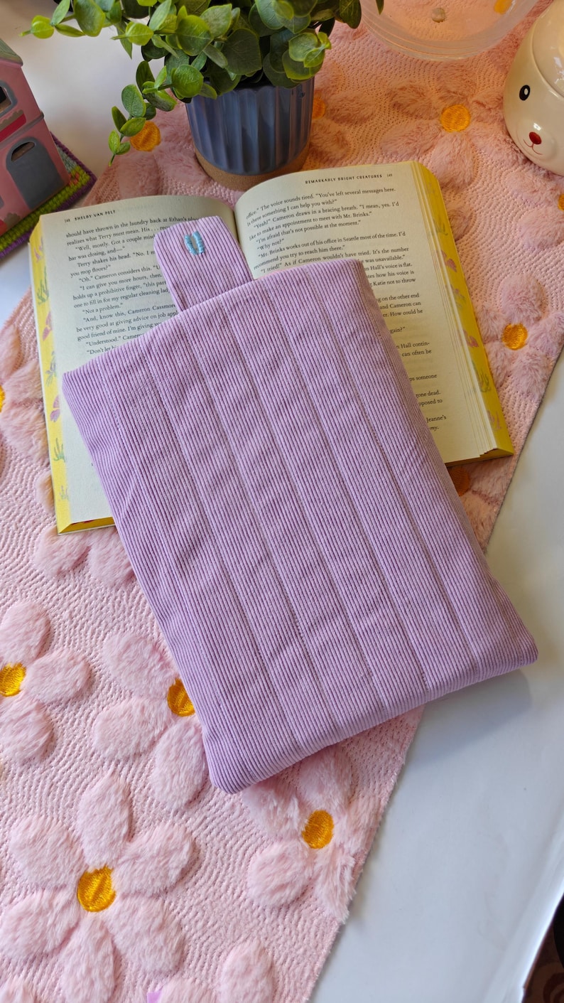 May include: A rectangular lavender corduroy pouch with a vertical ribbed texture and a small matching tab at the top center. The pouch is positioned flat on an open book, which rests on a soft, light pink fabric surface with a raised floral pattern.