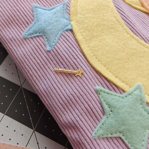 May include: A light purple corduroy fabric featuring stitched felt appliques of a large yellow crescent moon, a peach-colored sun, and several light blue and mint green stars. A small gold-toned metal shooting star charm is attached to the fabric.