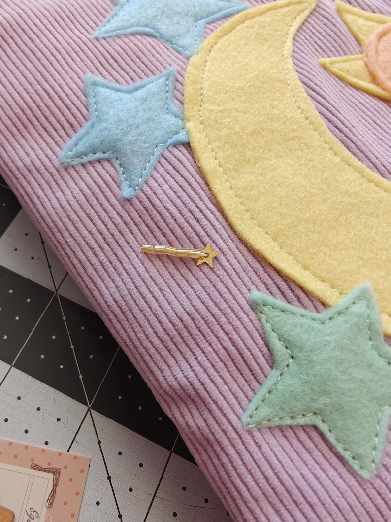 May include: A light purple corduroy fabric featuring stitched felt appliques of a large yellow crescent moon, a peach-colored sun, and several light blue and mint green stars. A small gold-toned metal shooting star charm is attached to the fabric.