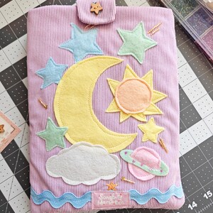 May include: A lavender corduroy fabric pouch featuring a felt applique design of a yellow crescent moon, a sun, a white cloud, a pink planet with rings, and several light blue and yellow stars. A small wooden star button secures the top flap.