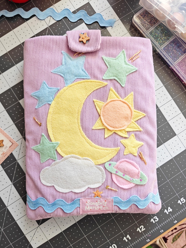 May include: A lavender corduroy fabric pouch featuring a felt applique design of a yellow crescent moon, a sun, a white cloud, a pink planet with rings, and several light blue and yellow stars. A small wooden star button secures the top flap.