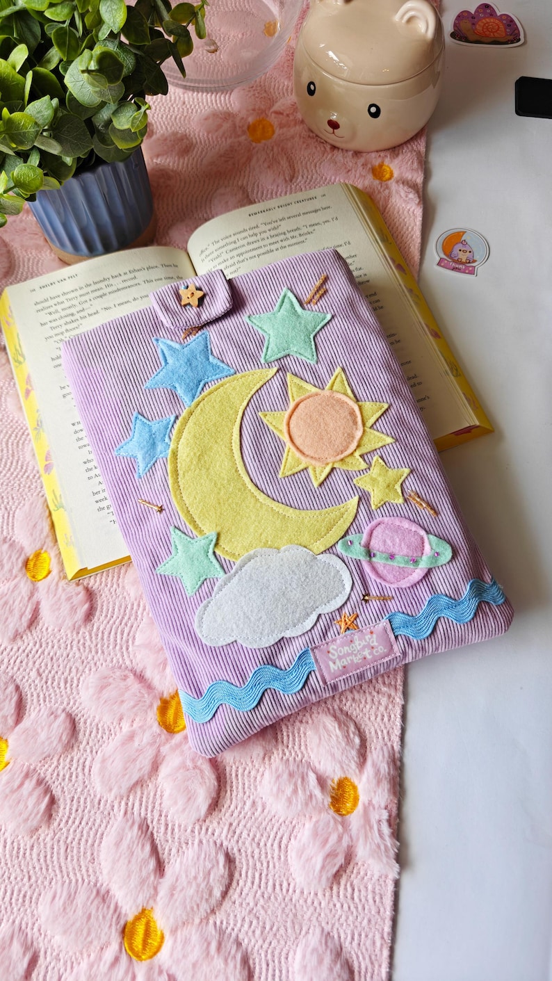 A handmade lavender corduroy book sleeve lies flat on a white surface, resting on a pink floral-patterned rug. The sleeve is decorated with colorful fabric appliqués of a large yellow crescent moon, a yellow and orange sun, a pink ringed planet, and several light blue and green stars. Small, hand-beaded gold details represent shimmering stars scattered across the design, and a wavy blue trim accents the bottom edge. A small wooden star-shaped button secures the top flap.