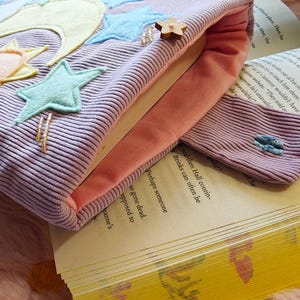 May include: A lavender corduroy book sleeve with a peach-colored interior lining. The exterior features felt appliqu&eacute;s of a yellow crescent moon, a small sun, and several light blue stars. A small wooden star button is attached to the top edge.