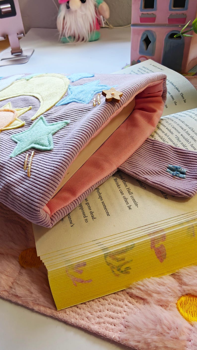 May include: A lavender corduroy book sleeve with a peach-colored interior lining. The exterior features felt appliqu&eacute;s of a yellow crescent moon, a small sun, and several light blue stars. A small wooden star button is attached to the top edge.