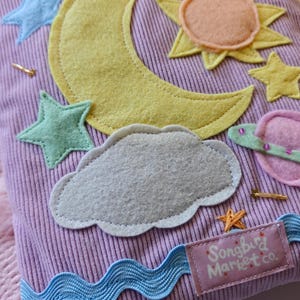 May include: A lavender corduroy fabric pouch decorated with felt appliqu&eacute;s of a yellow crescent moon, a peach and yellow sun, a gray cloud, a pink planet, and several light blue and yellow stars. A blue wavy ribbon and a label reading Songbird Market co.
