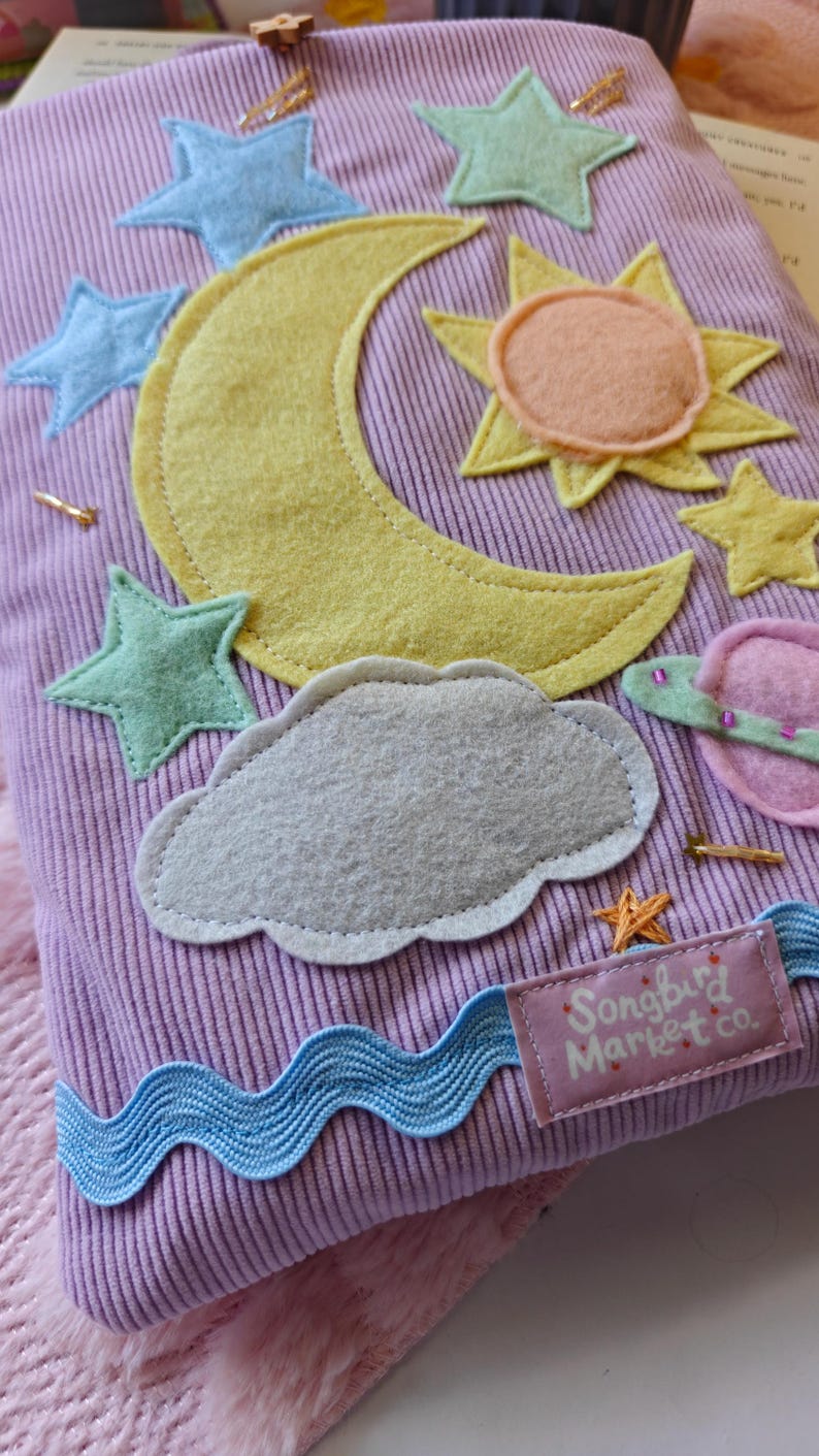 May include: A lavender corduroy fabric pouch decorated with felt appliqu&eacute;s of a yellow crescent moon, a peach and yellow sun, a gray cloud, a pink planet, and several light blue and yellow stars. A blue wavy ribbon and a label reading Songbird Market co.